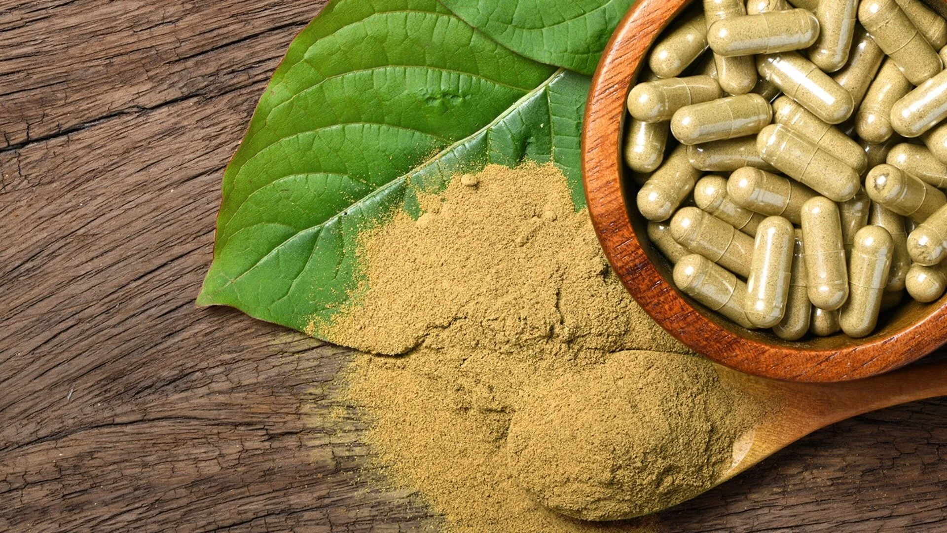 Kratom leaf with powdered kratom and capsules displayed on a wooden surface, highlighting Christopher's Organic Botanicals products.